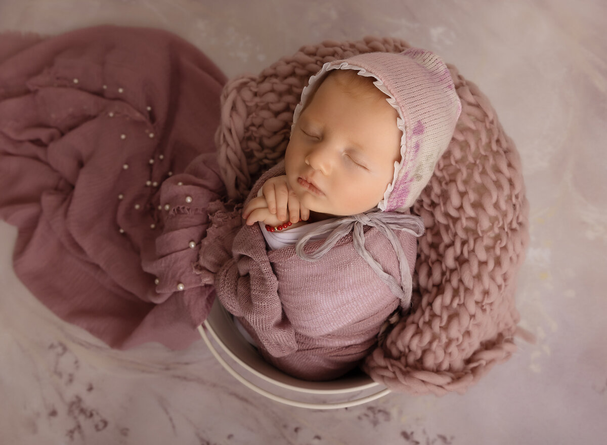 Aerial image of brooklyn newborn photoshoot. Baby girl is wrapped in lavender with her hands folded underneath her chin. Baby is placed in a bucket with mauve fabric and knits cascading beside her. Captured by top Brooklyn newborn photographer Chaya Bornstein