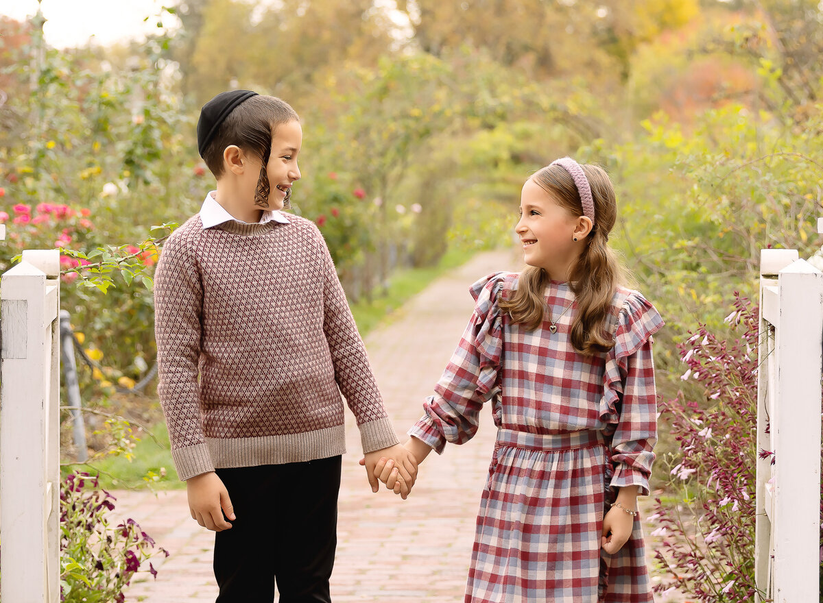 Two young siblings are captured at a Brooklyn, NY family photoshoot. The siblings are holding hands and looking at each other with a trail and wildflowers behind them. Captured by premier Brooklyn NY family photographer Chaya Bornstein Photography.