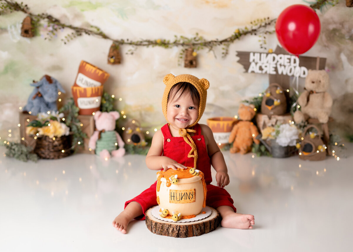 Winnie the Pooh themed cake smash in premiere West Palm Beach and Jupiter newborn cake smash and portrait photographer studio. Baby is wearing red overalls and a knit Winnie the Pooh hat with stuffed vintage characters and signage in the background. The baby is touching the top of a hunny pot cake.