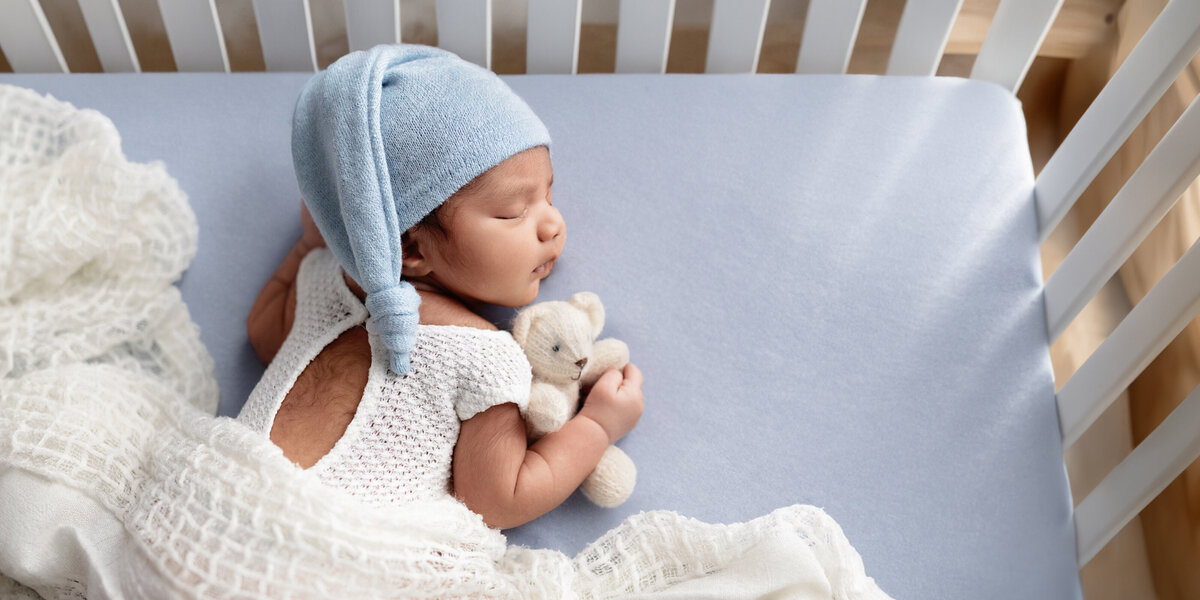 Newborn baby boy wearing a blue sleepy cap, resting on a blue crib mattress while holding a tiny knit teddy bear.
