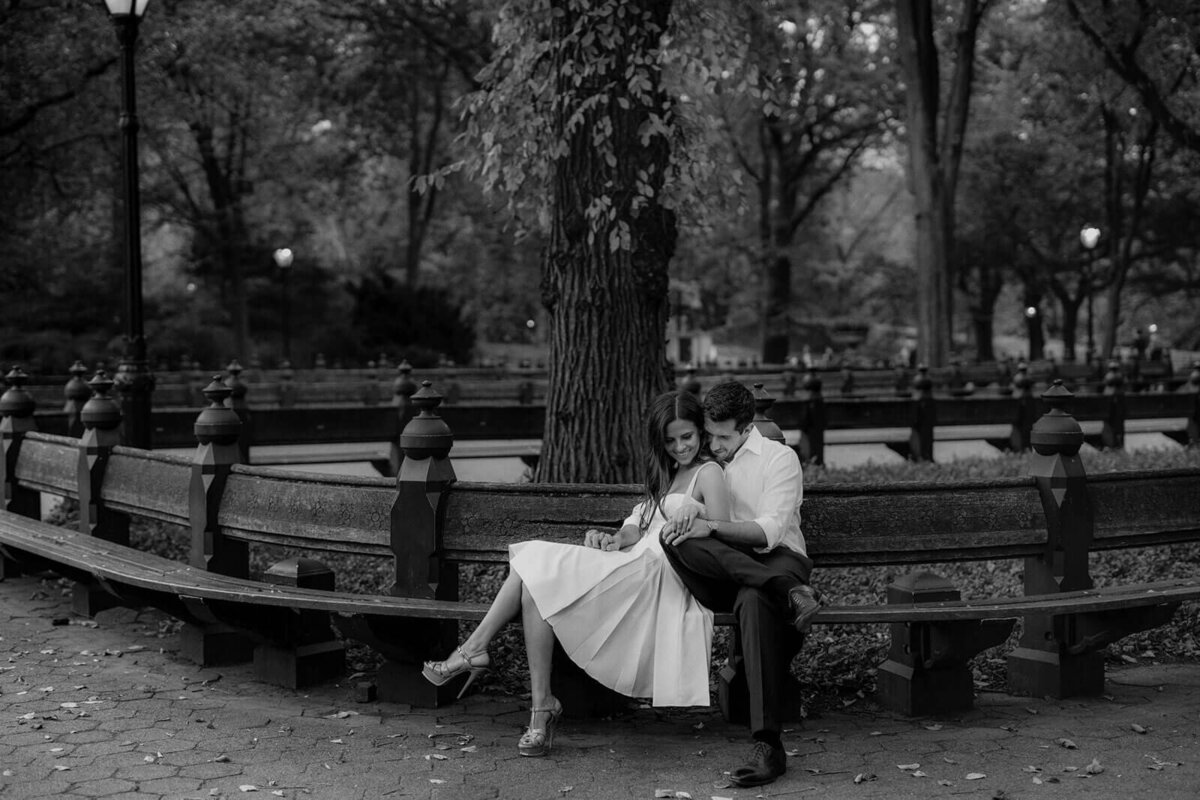 Black and white photo of the happily engaged couple sitting on the bench at Central Park, NYC. Image by Jenny Fu Studio.