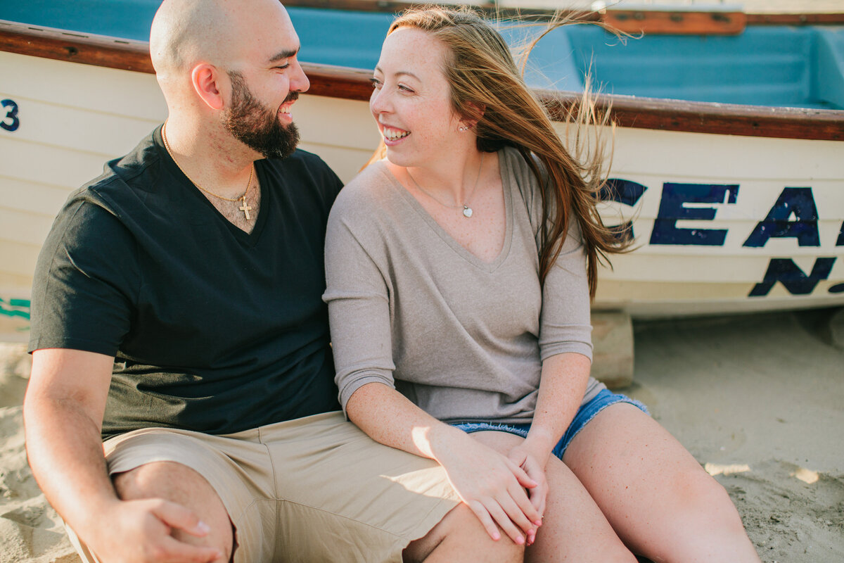 A couple sitting on the ground in front of a row boat smiling at each other 