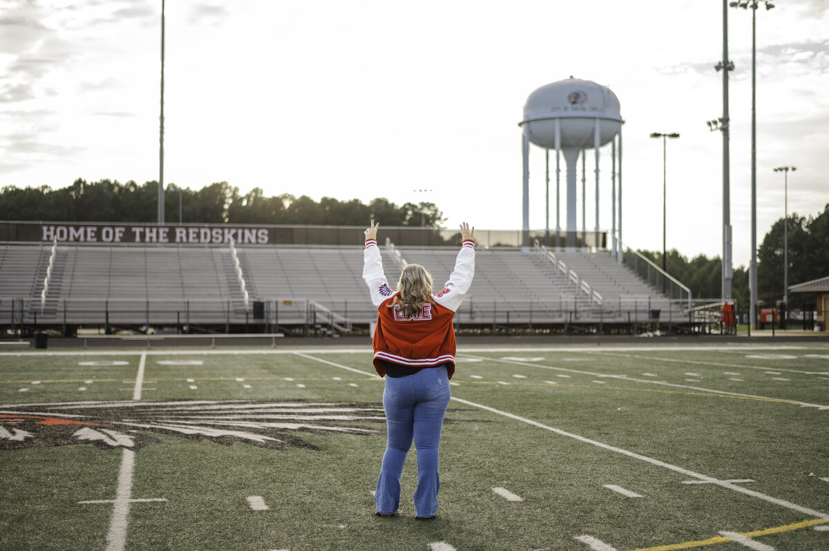 High school senior wearing letterman jacket stands in the middle of football field with hands in the air. Photo taken by Atlanta senior photographer Amanda Touchstone