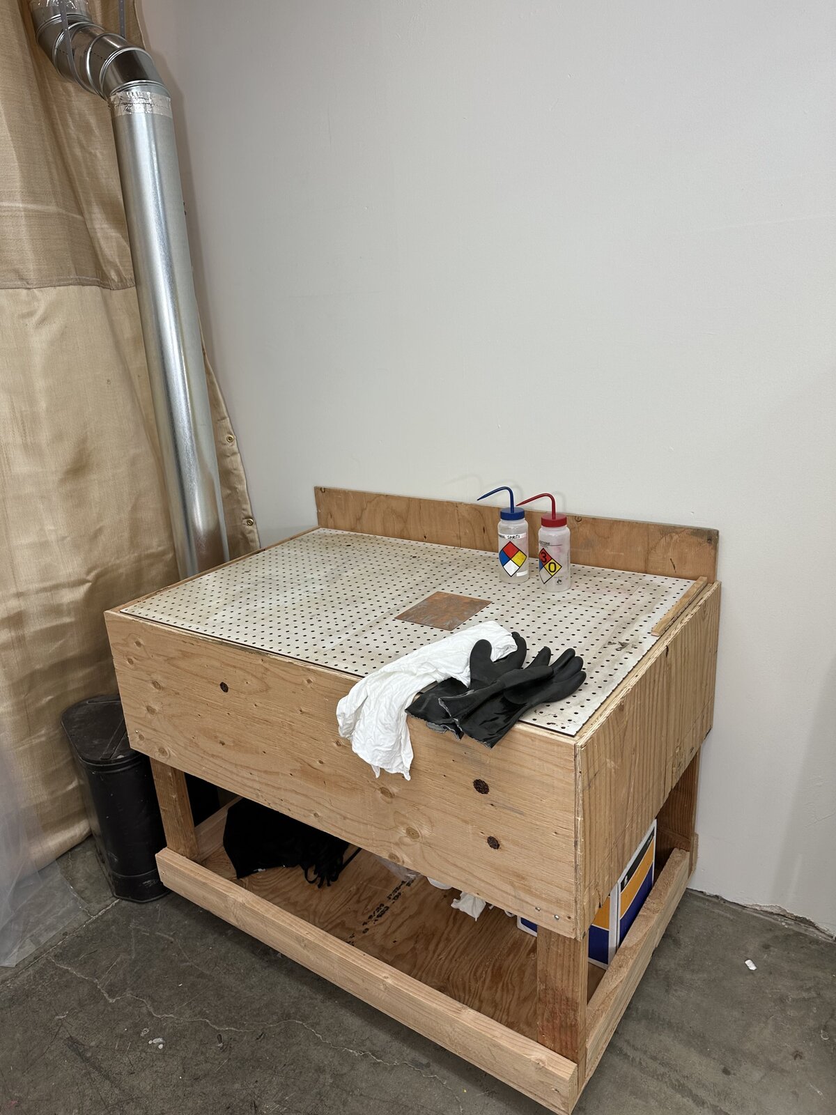 A corner of a printmaking studio with a ventilation system and a sturdy workbench, equipped with safety gloves and cleaning supplies for printmaking processes.