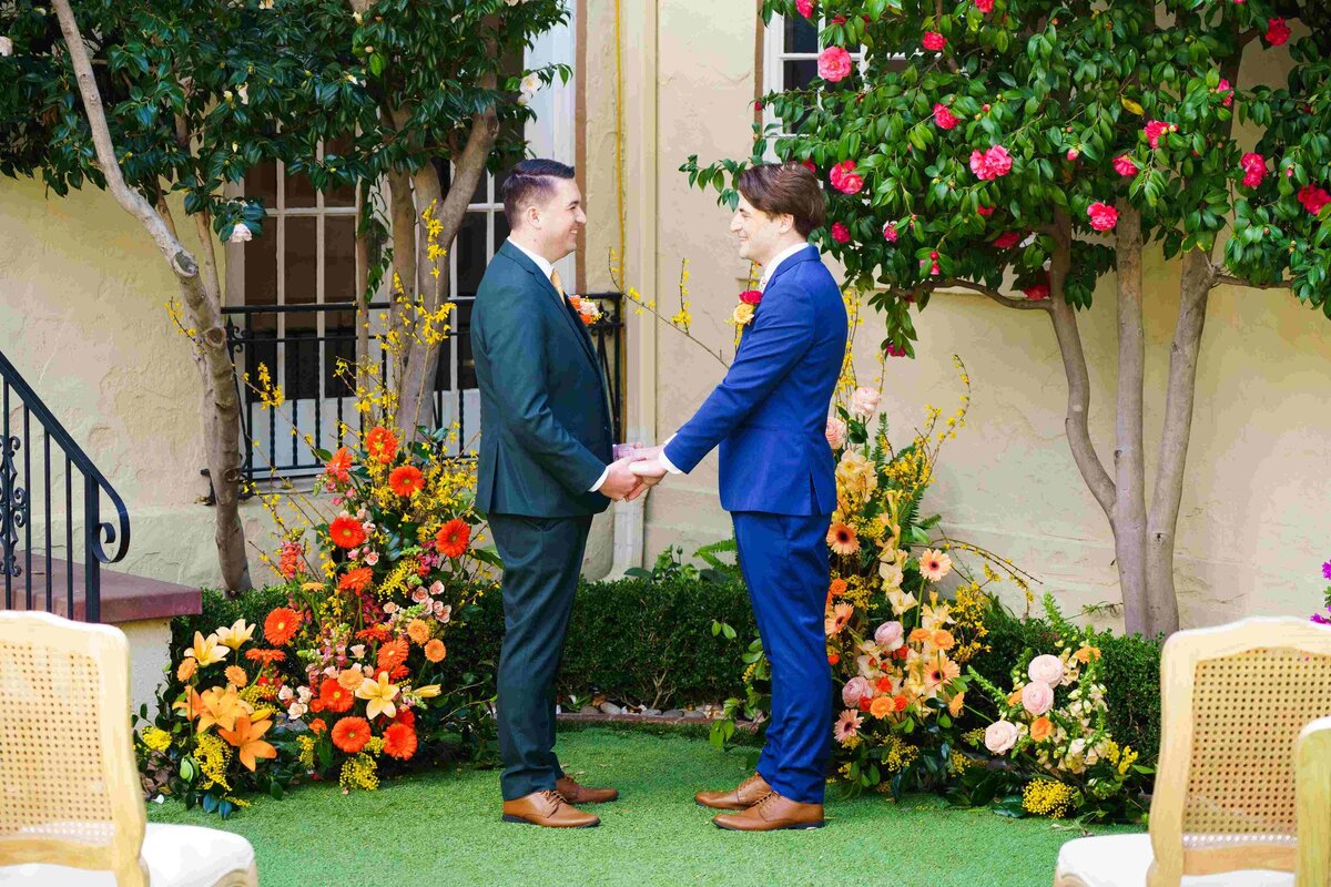 Two grooms holding hands in blue and green suits at their wedding ceremony