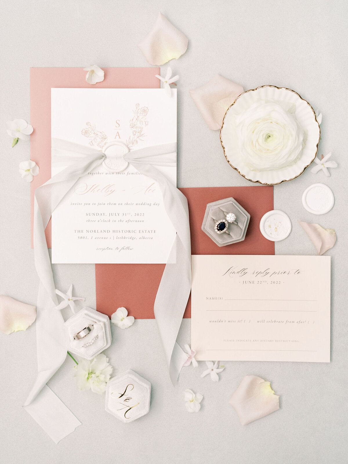 An elegant wedding invitation set for a destination wedding in Canada includes a main card, RSVP card, white ribbon, rings in boxes, a white flower, and scattered petals, arranged against a light background with a pink envelope. Imagine this beautiful setup for your Banff Alberta wedding.