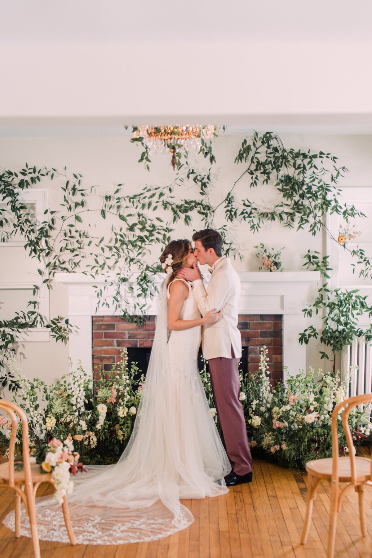 Closeup of bride wearing Alexandra Grecco kissing groom in white tuxedo surrounded by vines and flowers in wedding ceremony