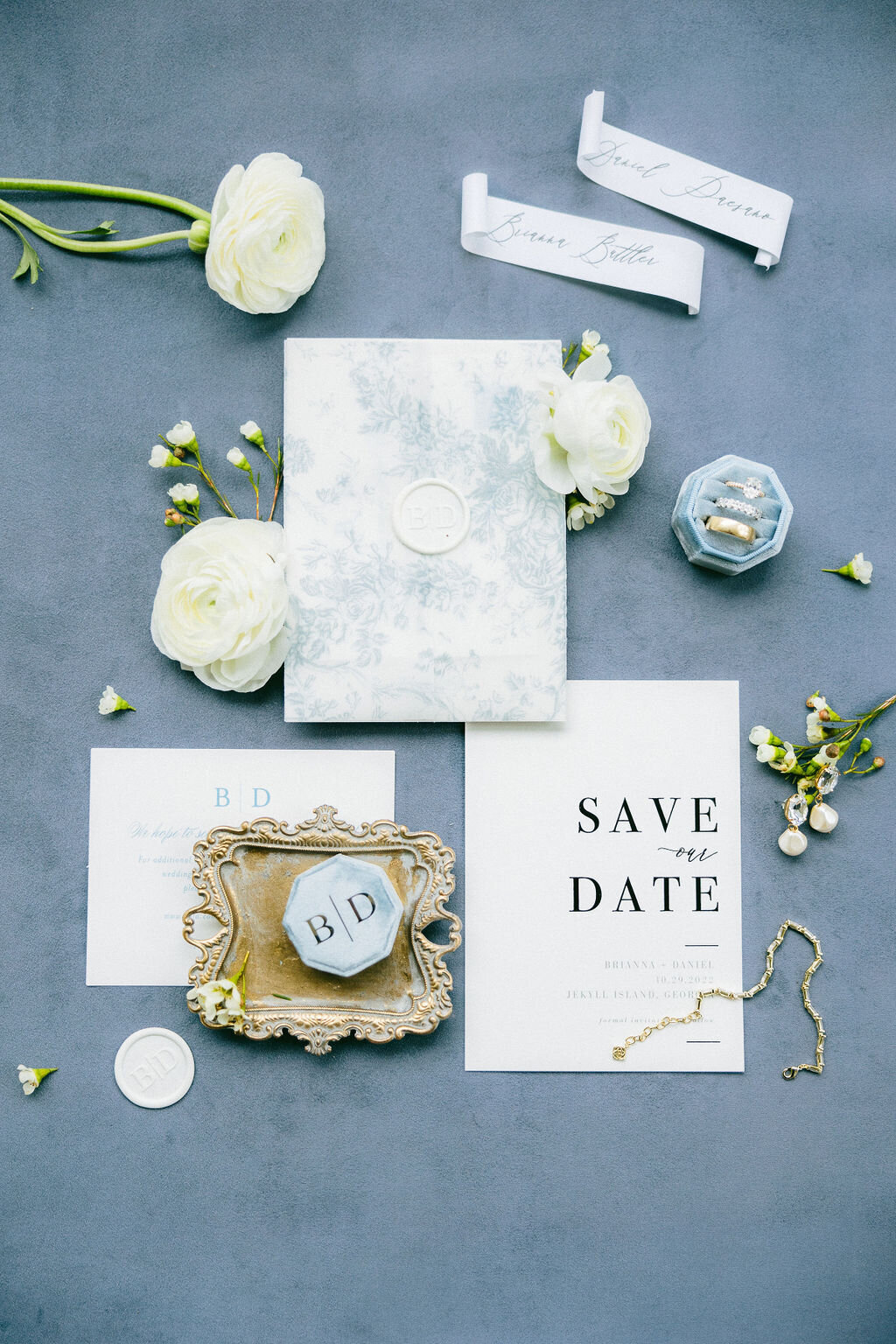 Blue-toned “Save the Date” suite with velvet ring box, wax seals, and white ranunculus—styled flat-lay for a Sea Island wedding in South Carolina, captured by luxury wedding photographer Amia Marcell.