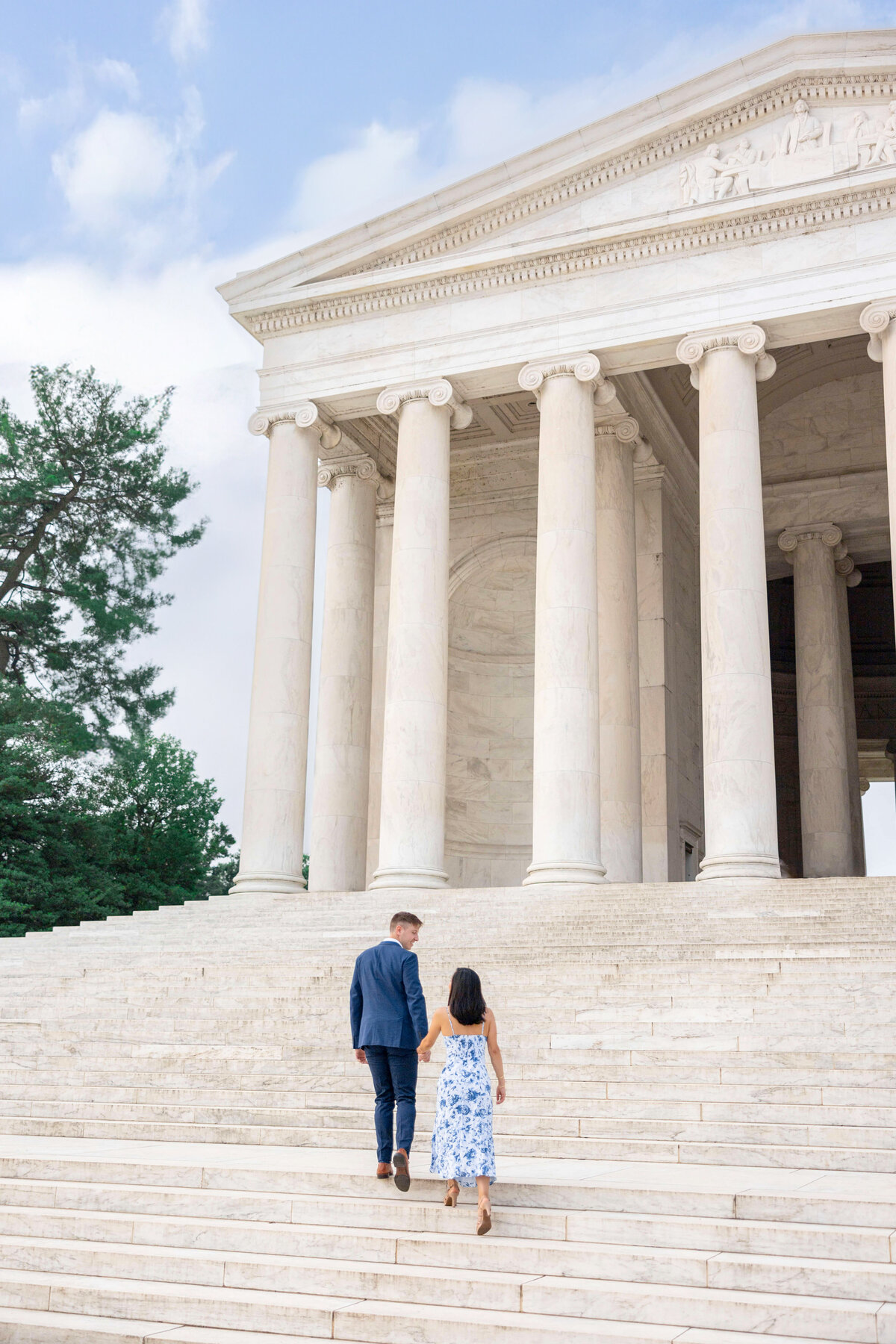 A couple walks hand in hand up the marble steps of a neoclassical building with large columns, set against a blue sky with scattered clouds. A tree is visible on the left side of the building.