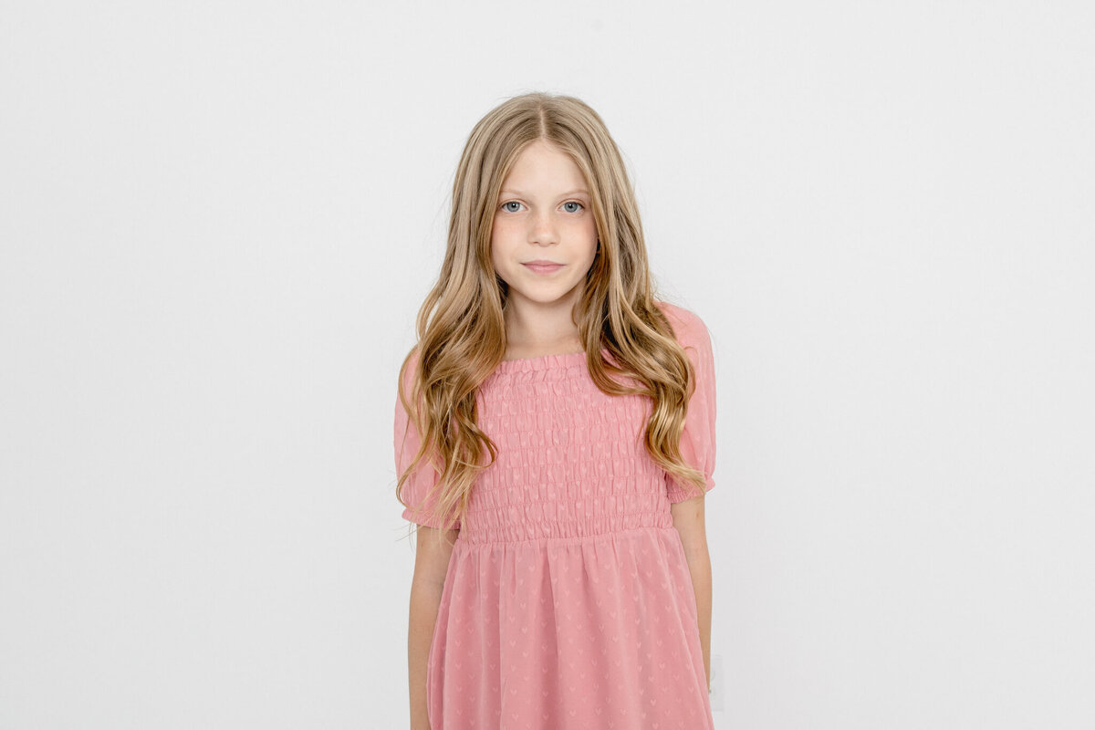 A young girl with long, blonde hair wearing a pink dress smiling with her lips and looking at the camera against a white background in Tustin.