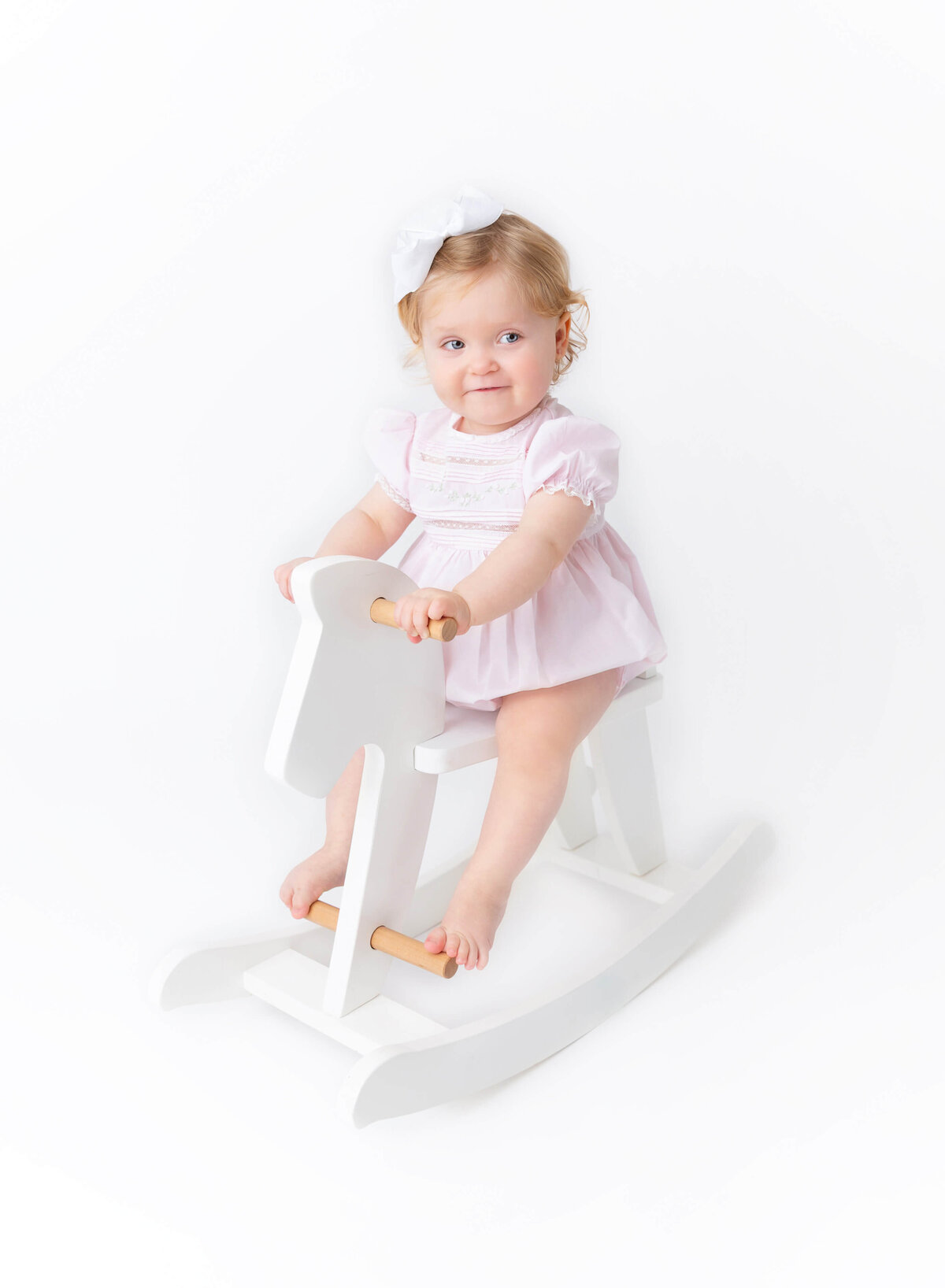Baby girl sitting on a white wooden rocking horse in a classic pink outfit — timeless modern Heirloom baby portrait captured by NYC Heirloom photographer in Brooklyn studio.