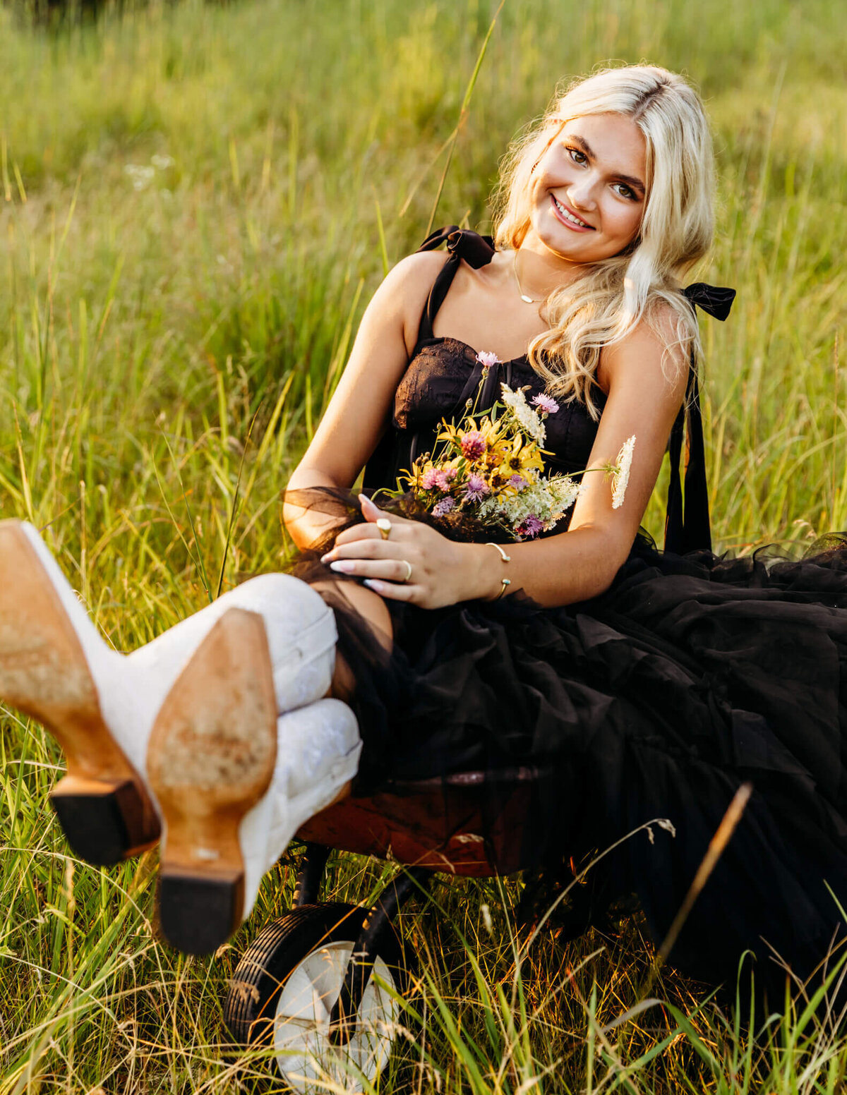 teen girl in a black dress holding a bouquet of flowers as she sits in a wheelbarrow with her feet crossed in the middle of a field in Neenah.