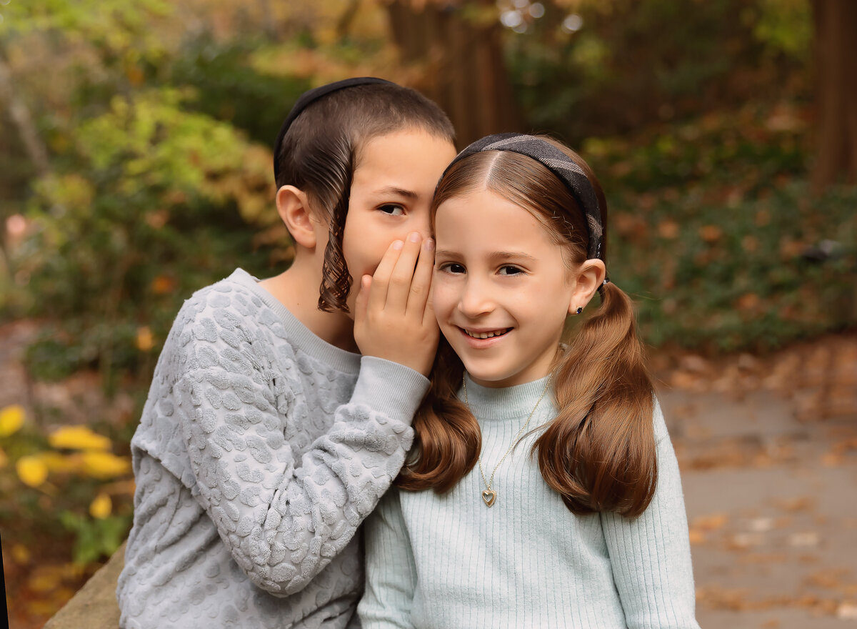 Big brother is whispering a secret to his little sister at a fall family photoshoot. Captured by Brooklyn family photographer Chaya Bornstein