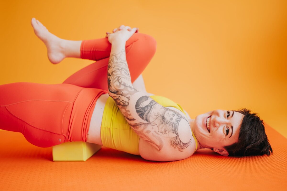 Woman with short brown hair smiles to the camera while wearing a yellow sports top and orange leggings. She stretches one folded leg into her chest in front of an orange backdrop.