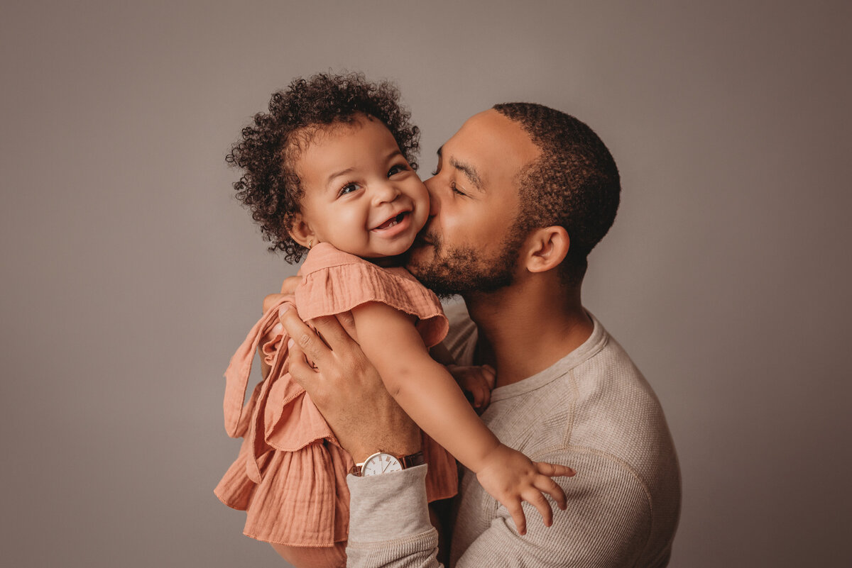 Family portrait on light grey backdrop with dad holding one year old baby girl kissing her cheek and baby is smiling at camera, wearing nude pink and creams