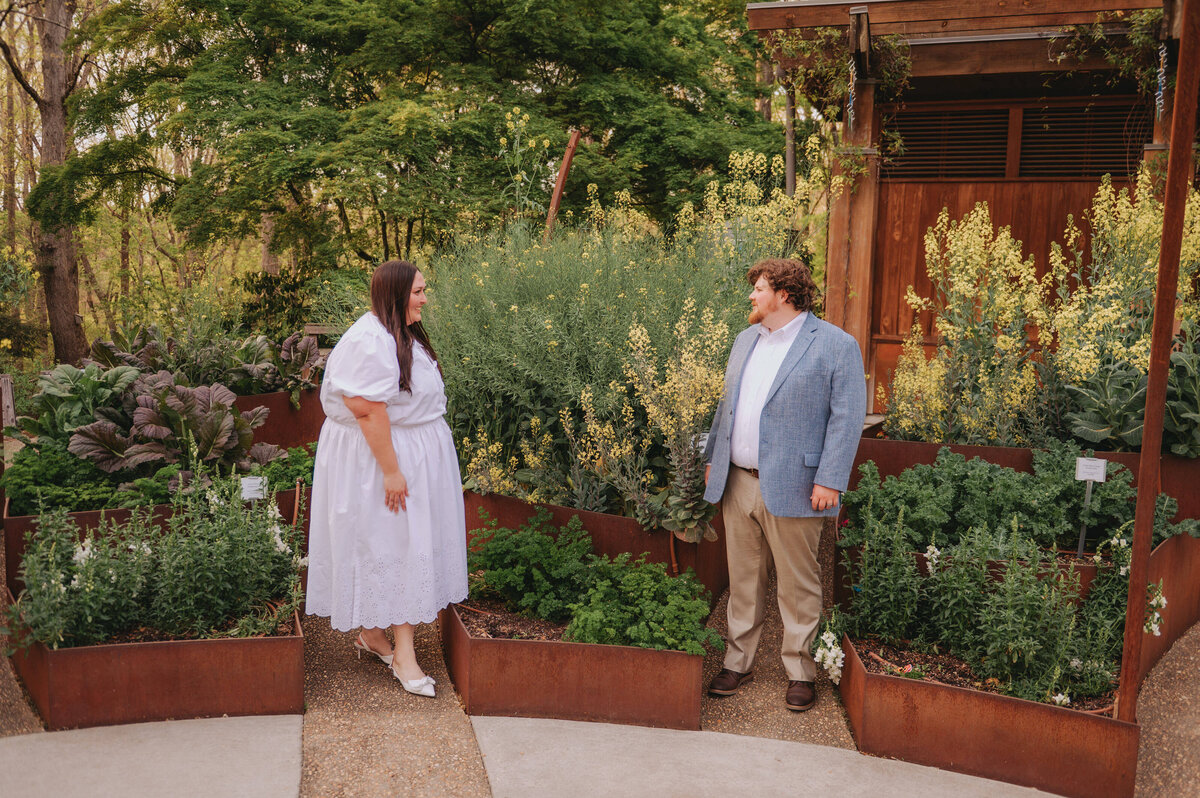 A couple stands outdoors, looking at each other, surrounded by greenery