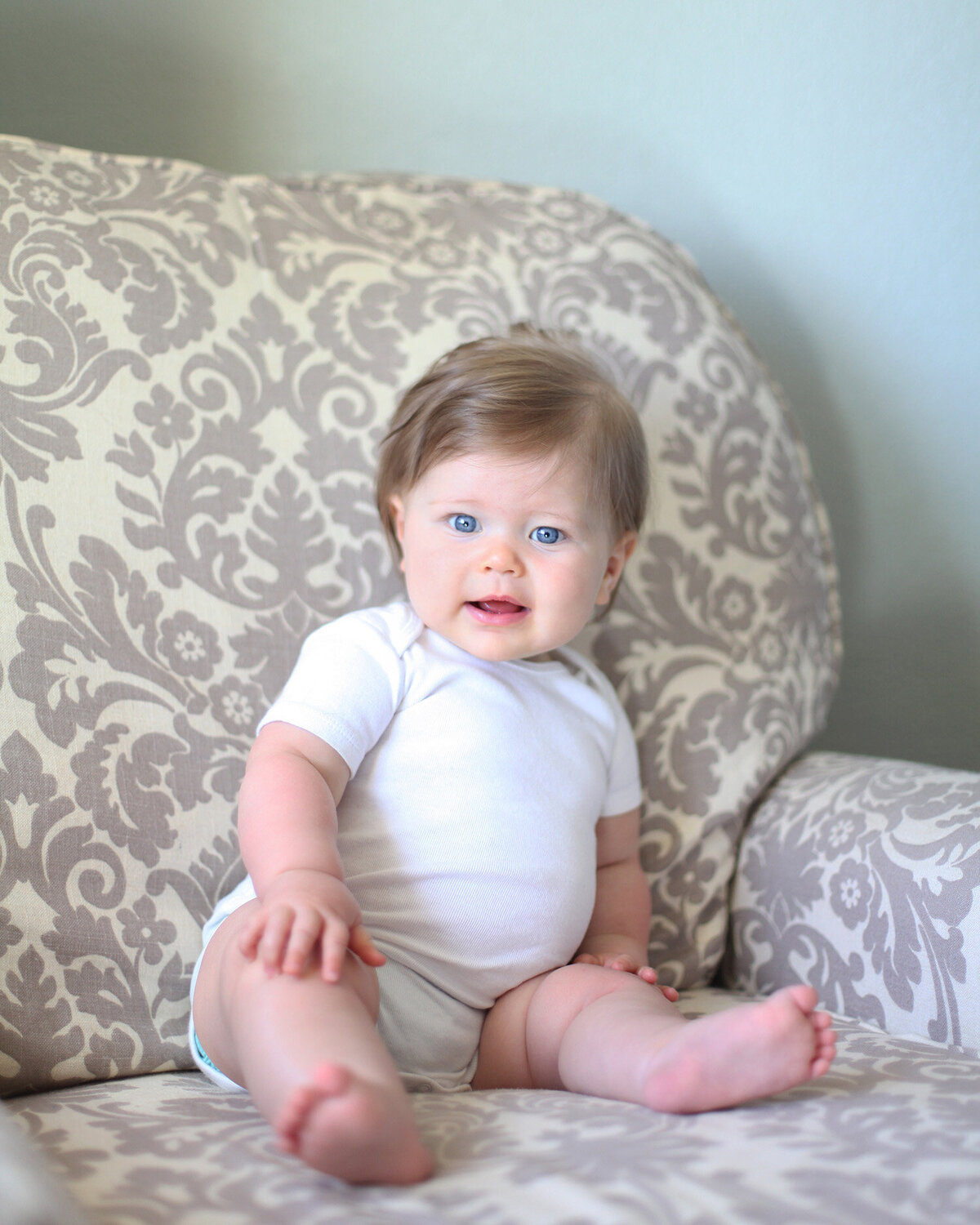Seven month old baby wearing a white onsie sitting on a couch lit by natural light looking at the camera captured at home by San Francisco Newborn Photographer.