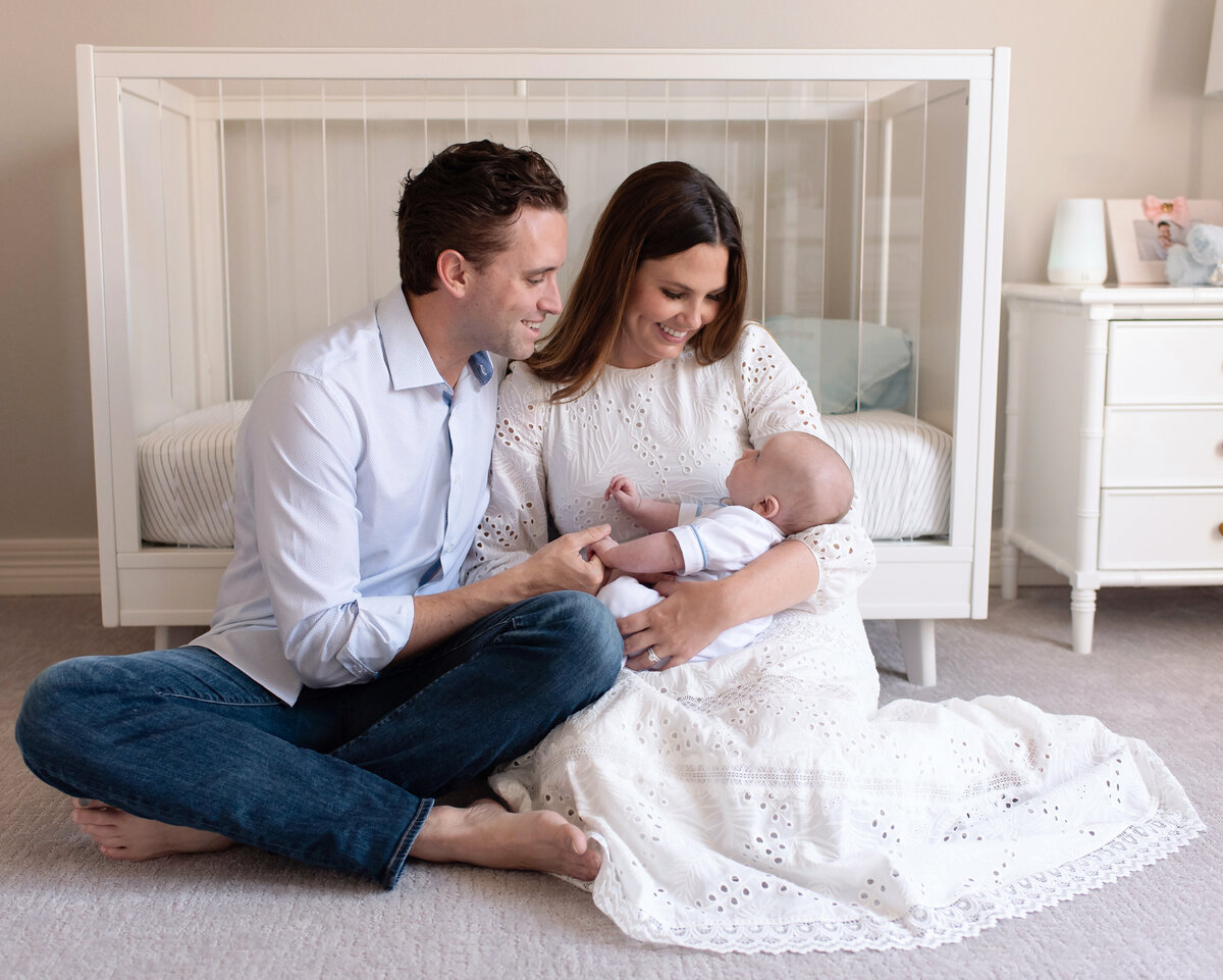 Parents sitting in front of a white crib in a baby nursery while playing with their newborn boy.