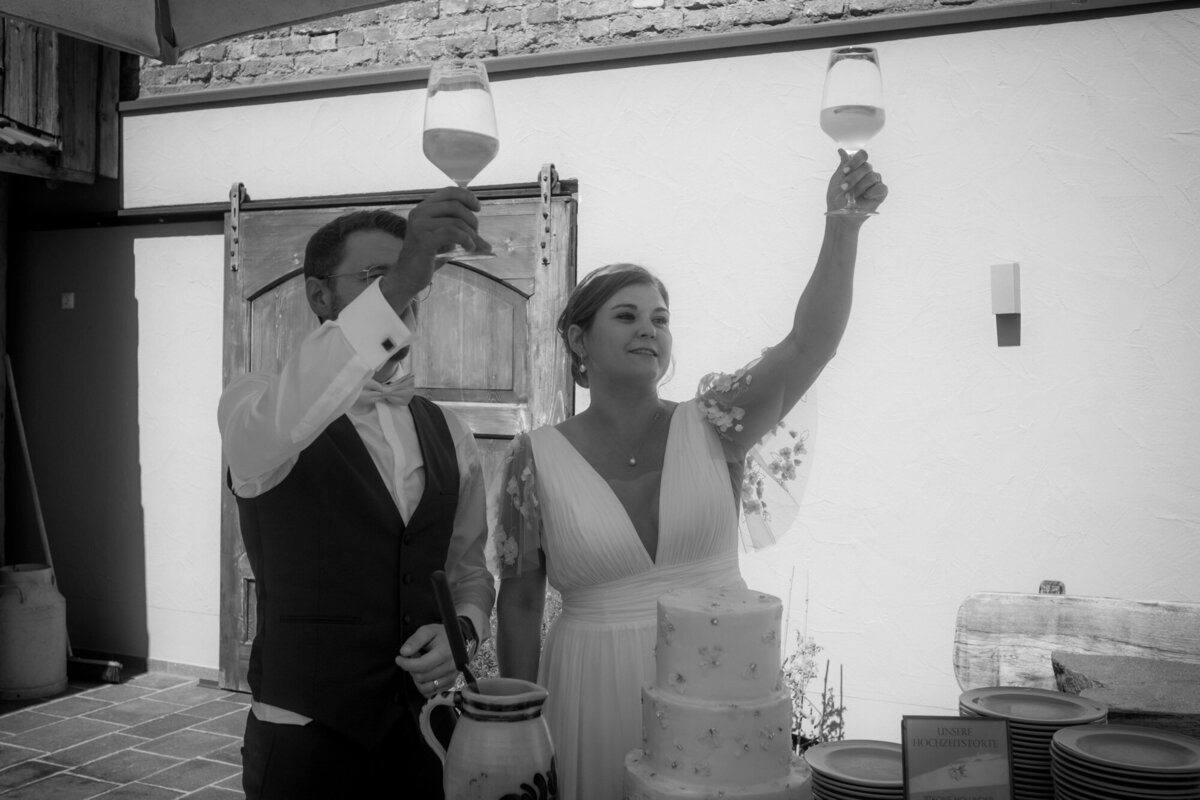 Das Hochzeitspaar steht hinter der Torte und prostet den Gästen mit erhobenen Gläsern zu.