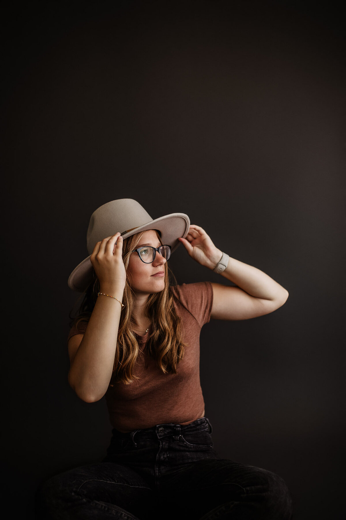 High school senior girl in dark and moody studio setup wearing glasses holding hat looking at the light