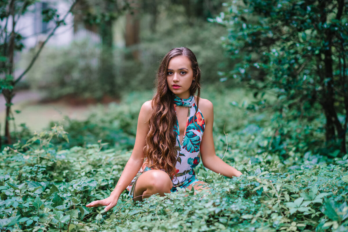Senior girl in tropical floral romper sitting in lush greenery during her style-focused photo session in Charleston, South Carolina