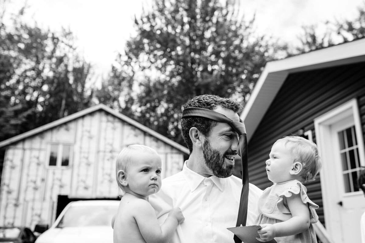 Dad wearing a tie on his head to the side, holding two babies—one his own—before his brother’s wedding in North Hatley, Quebec, captured in a playful black-and-white photo