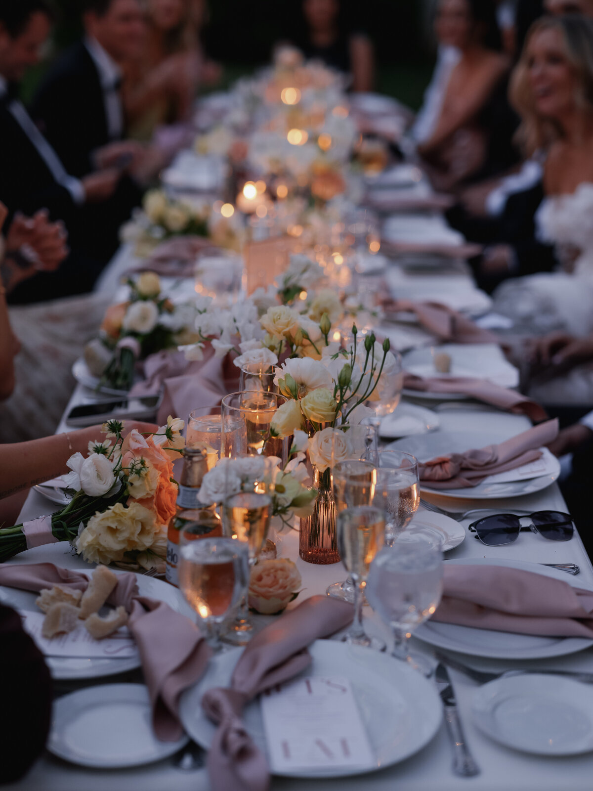 guests sat at outdoor wedding reception table with blush napkins, florals, wine glasses
