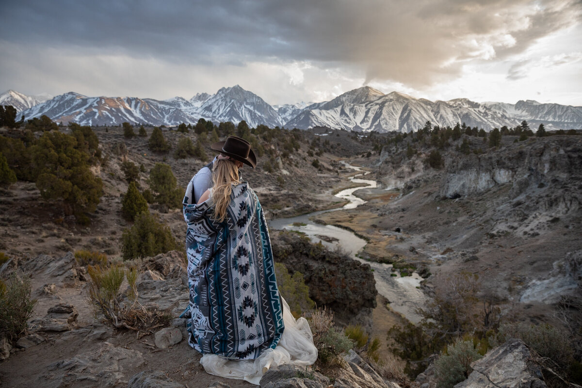 A bride and groom stand cuddling together under a blanket as the sunsets on their california elopement day.