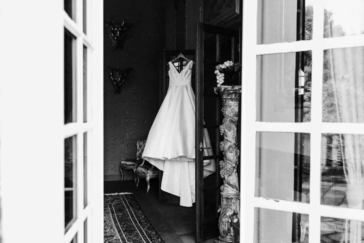 Jurk van bruid hangt in hotelkamer met openslaande deuren fotograaf fotografeert van buiten naar binnen zwartwit