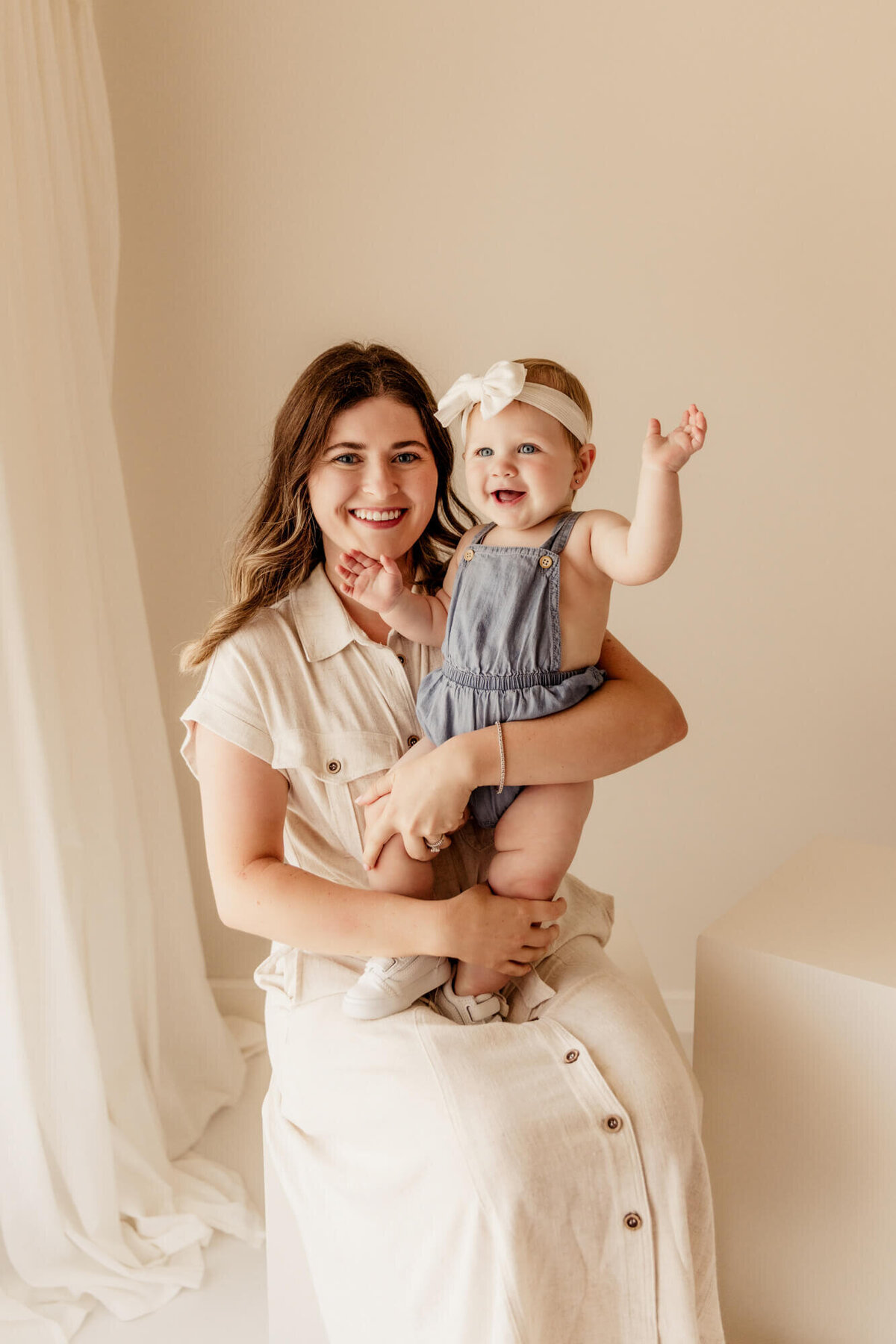 Mother wearing a cream dress and daughter in blue overalls smile for a portrait near Edmond Oklahoma.