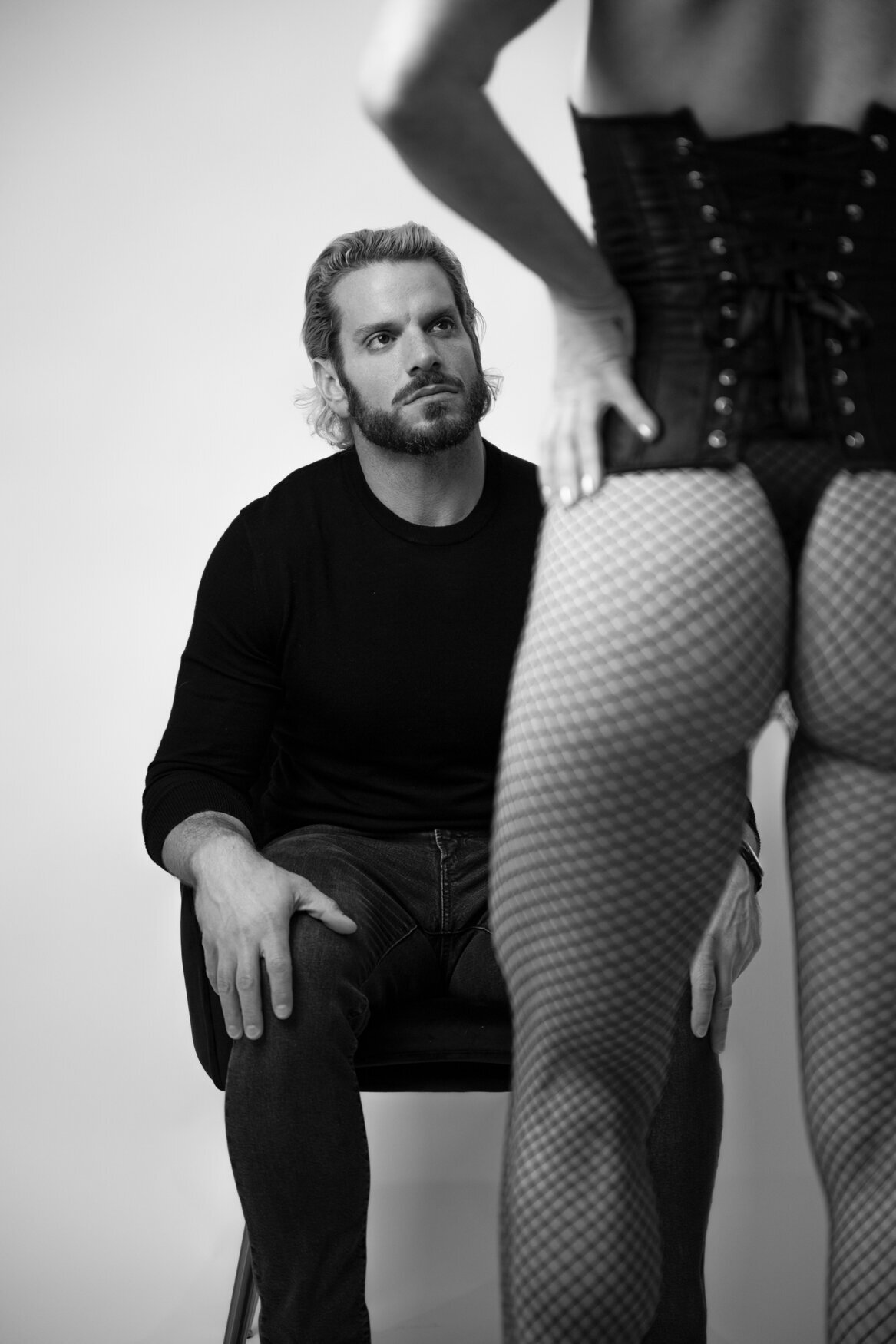 Black and white photograph of a couple in an intimate pose. The man, seated in a chair, gazes up at her while holding her waist gently. The scene exudes confidence, connection, and romance.