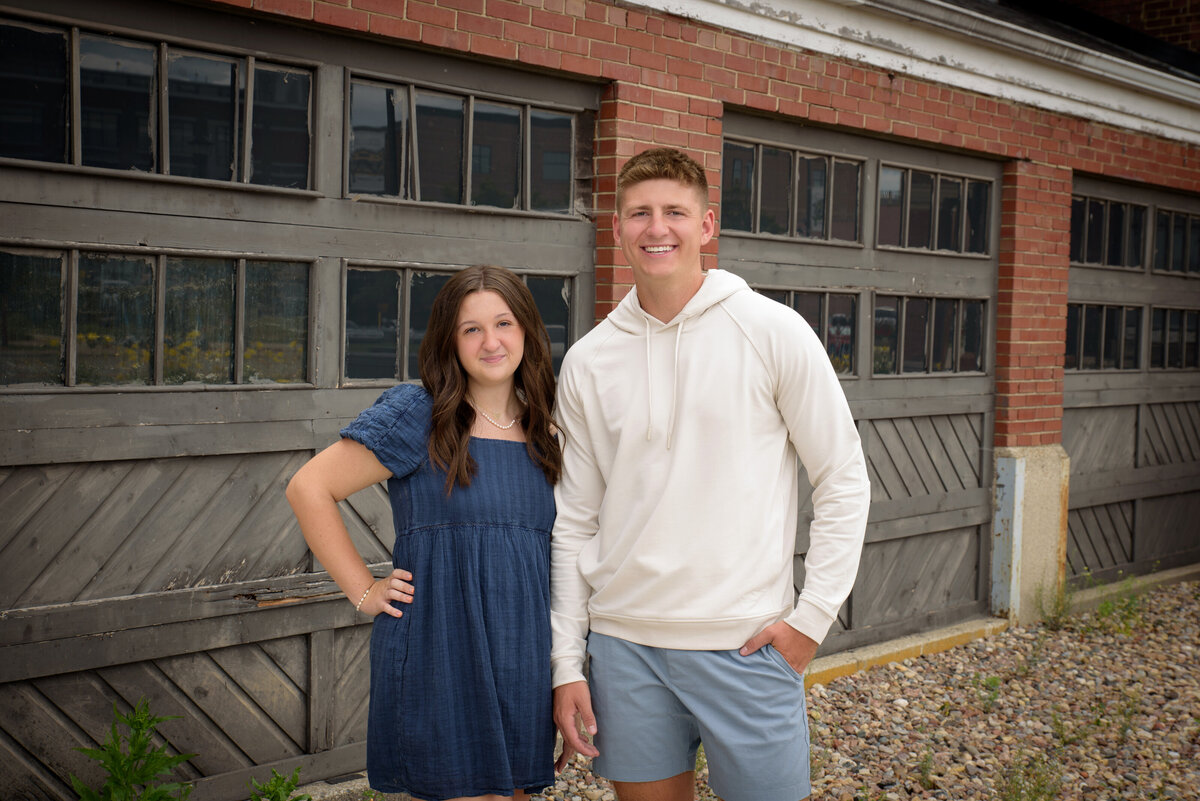 De Pere High School senior boy wearing blue shorts and a cream hooded sweatshirt with his sister for a buddy shot by The Depot in Downtown Green Bay, Wisconsin