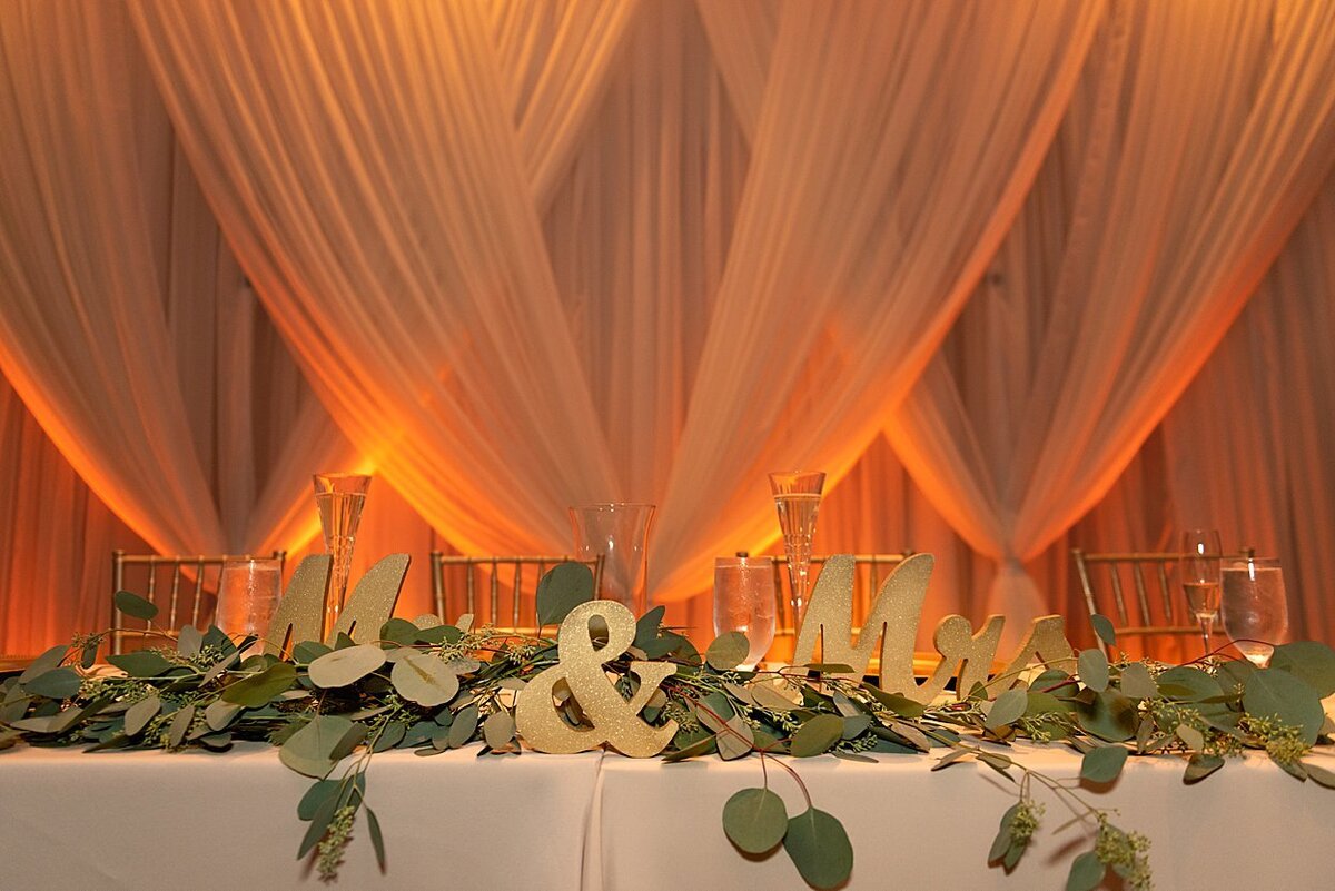 Photo of Bridal Table decorated with gold Mr. and Mrs and greenery with lit peach colored drapes in background