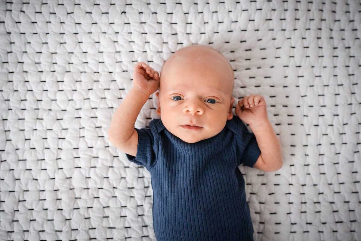 A close-up of a newborn baby lying on a textured white blanket, gazing up with wide eyes.