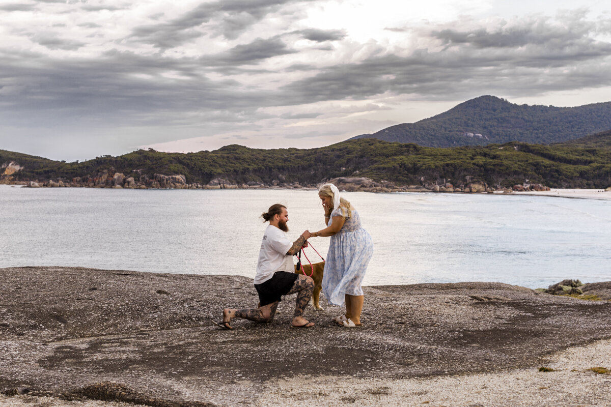 Bride and Groom standing on a mountain top. The wind is blowing in her hair and they are looking out to the view while hugging each other. The bride is a long sleeved wedding gown.