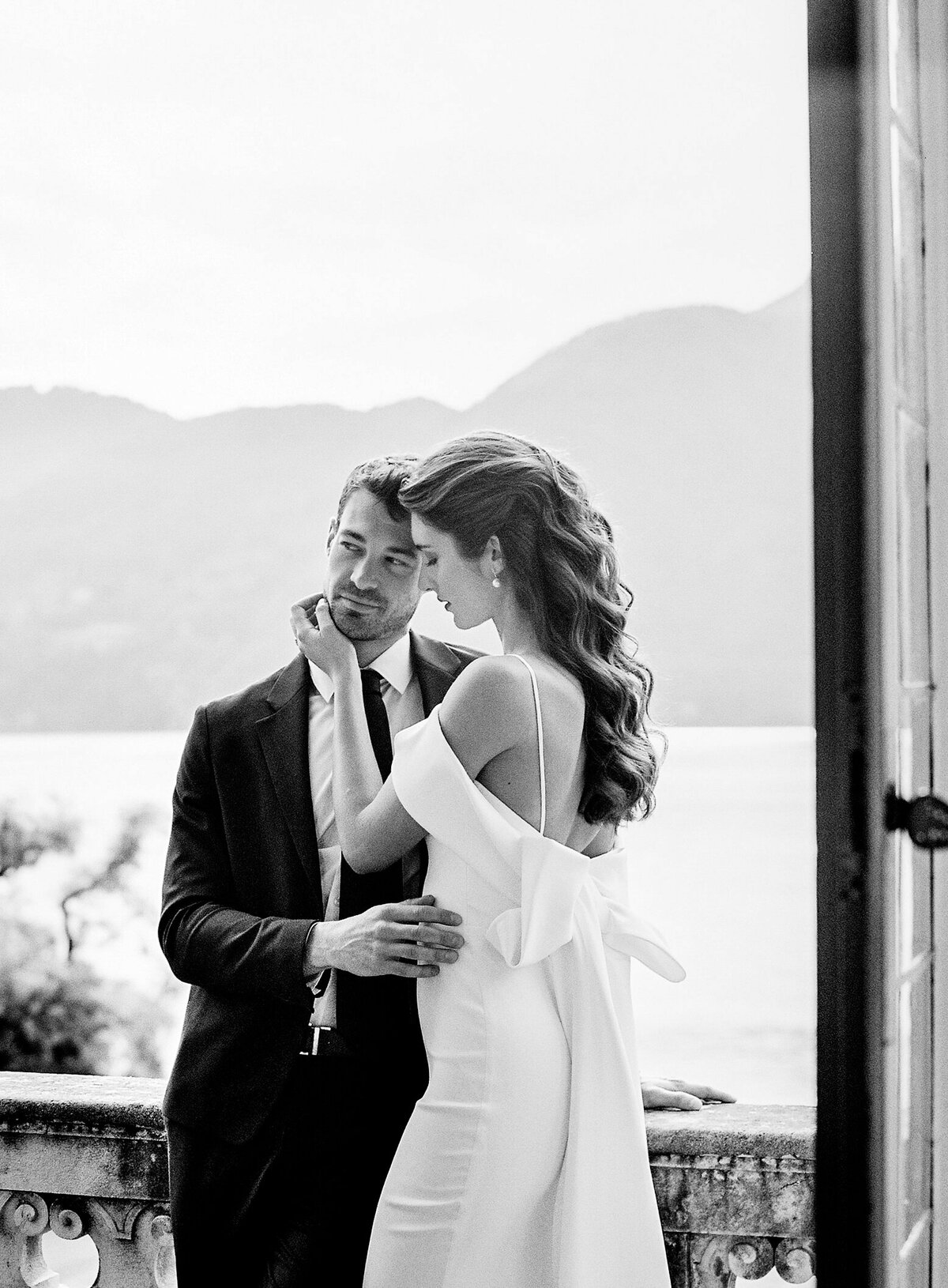 Black and white photograph of bride and groom on the balcony of Villa Sola Cabiati on Lake Como with the lake and the mountains in the background the bride is in an off the shoulder wedding gown with oversized bow in the back with groom in dark suit for their Lake Como wedding photographed by Italy wedding photographer