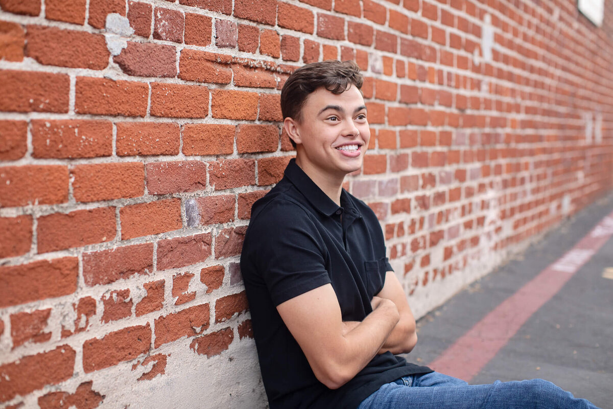 A person in a black shirt and jeans sits with arms crossed, leaning against a brick wall. They are smiling and looking off to the side. The ground is paved.