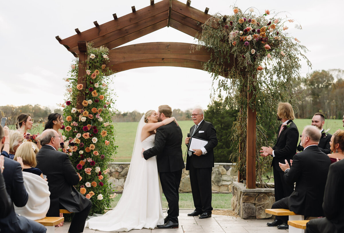 Charlottesville wedding photographer photographs bride adn groom sharing a passionate kiss at their outdoor ceremony with their guests clapping for them in celebration with green hills in the distance