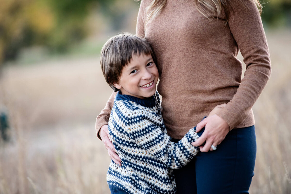 A boy in a stripe sweater hugs onto mom while standing in a field