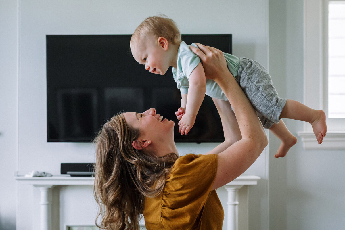 mom holding toddler son up above her head as both smile in front of tv at home