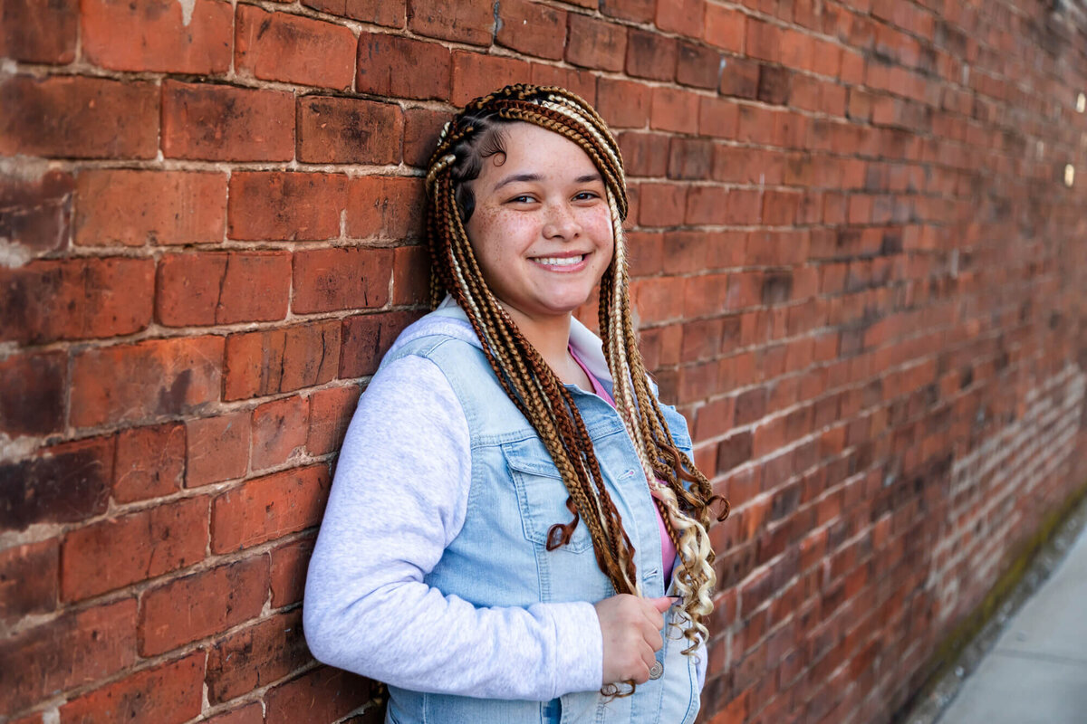 A lovely senior picture of a girl wearing braids and a denim hoodie leaning on a city brick wall. Captured by Springfield, MO senior photographer Dynae Levingston.