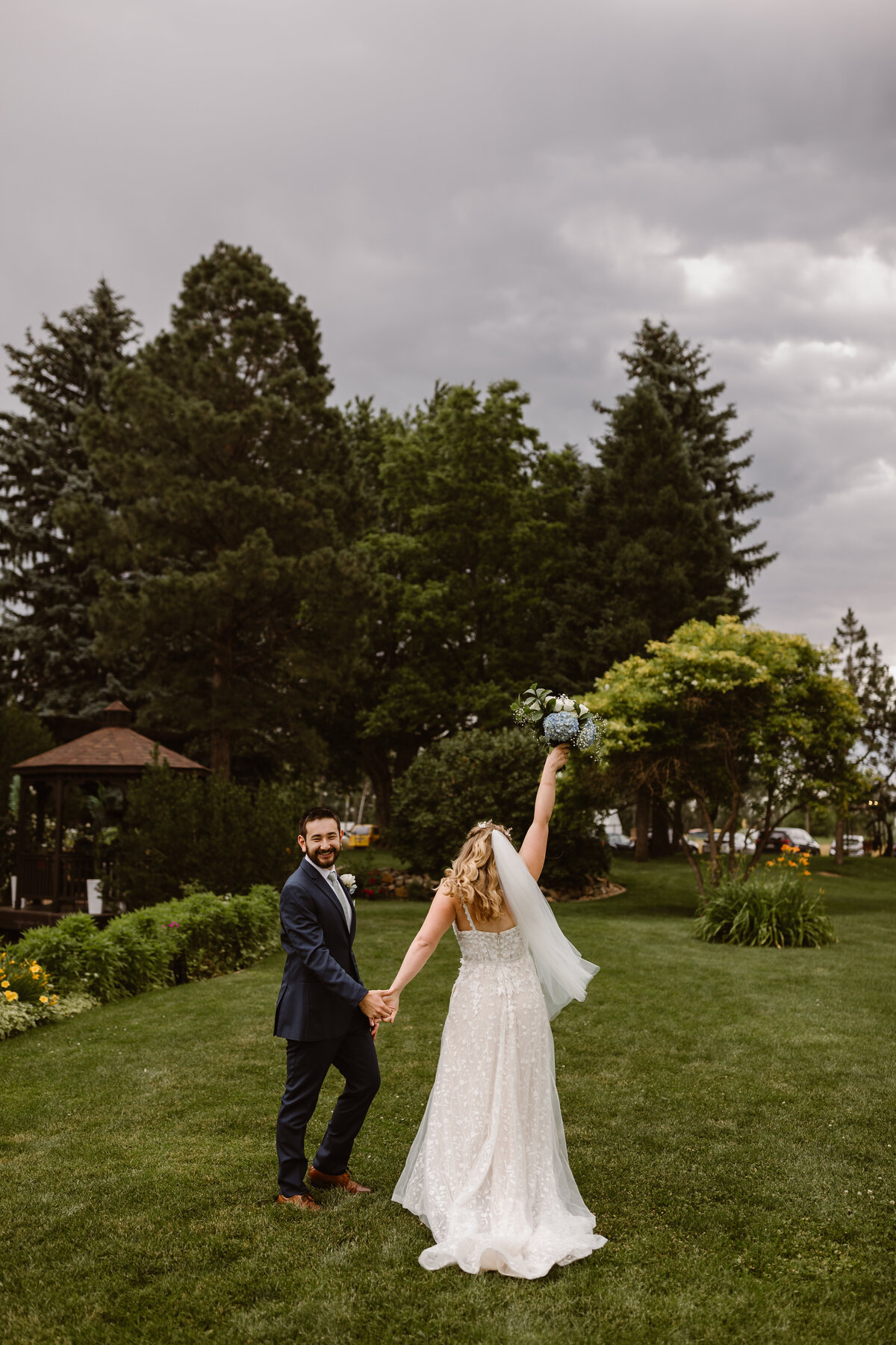 Bride and groom walking hand-in-hand across the lawn at Shupe Homestead, with the bride raising her bouquet in celebration and the groom smiling back at the camera.