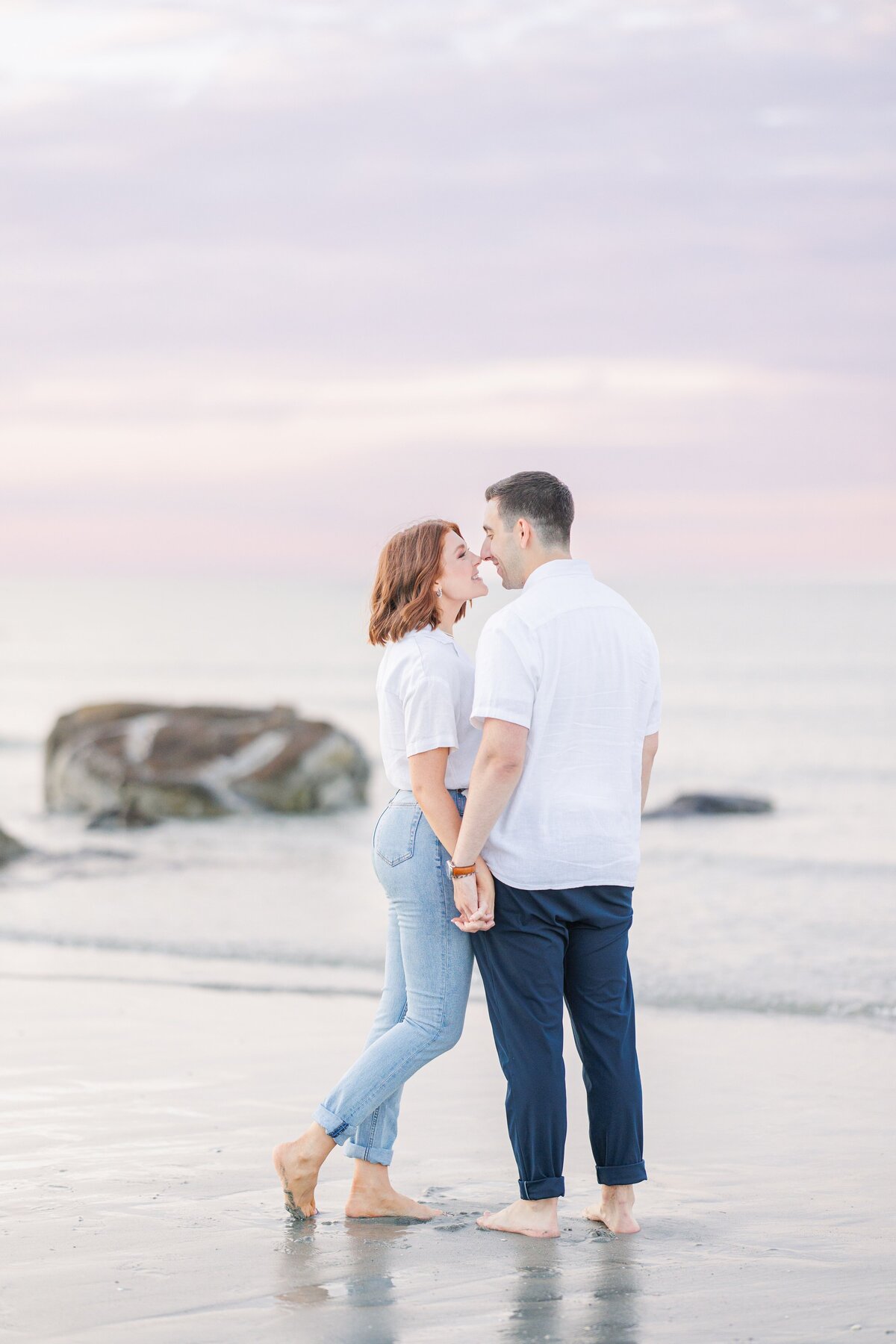 A man and woman smiling and almost kissing while holding hands during their Wingaersheek Beach engagement.
