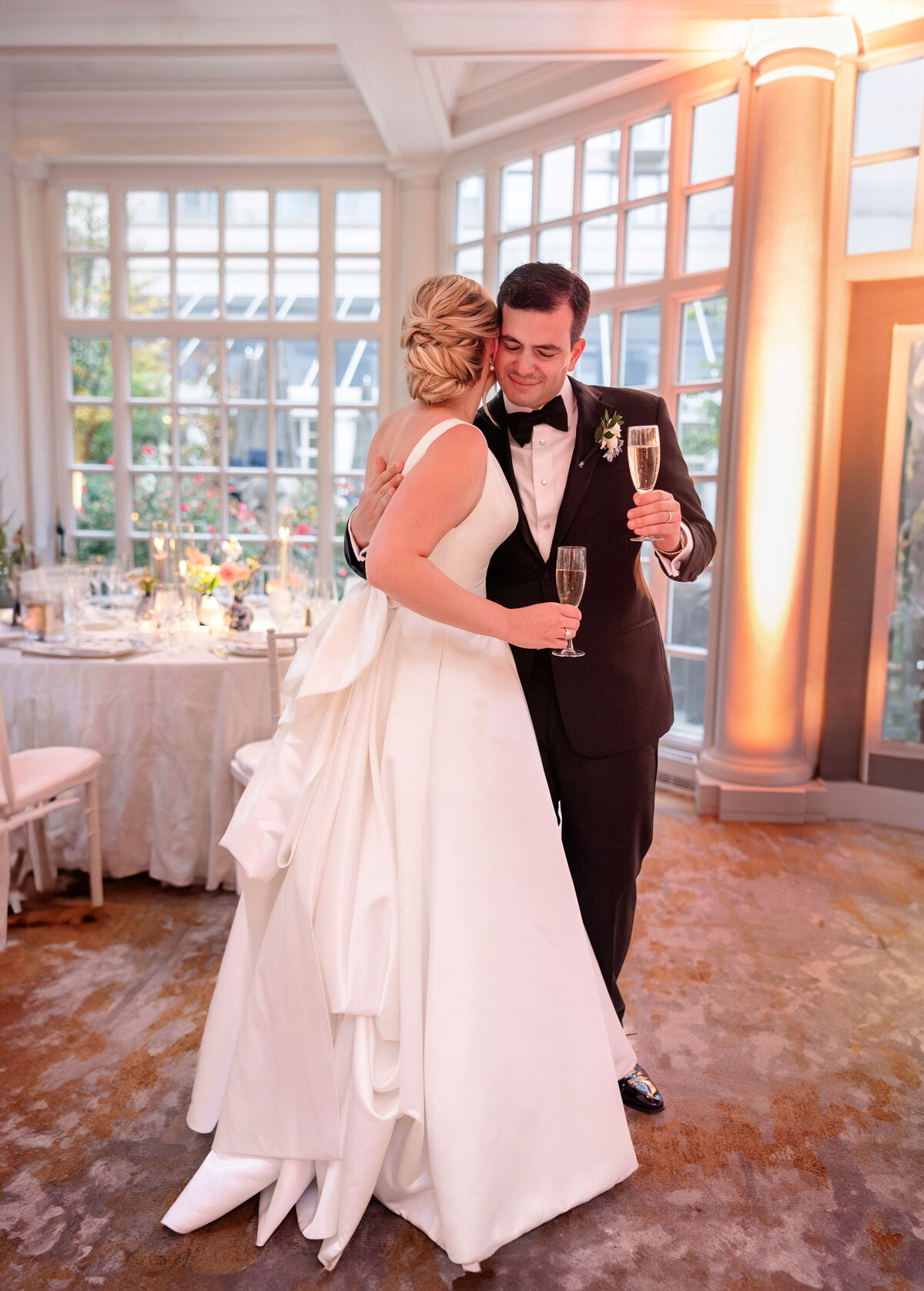 A bride and groom embrace while holding champagne glasses. The bride is wearing a white gown with a long train, and the groom is in a black tuxedo. They are in a bright room with large windows and elegant decor.