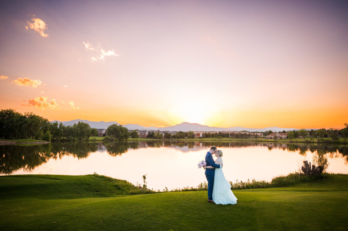 A wide angle shot of a bride and groom standing forehead to forehead in front of a lake at The Barn at Raccoon Creek in Colorado.