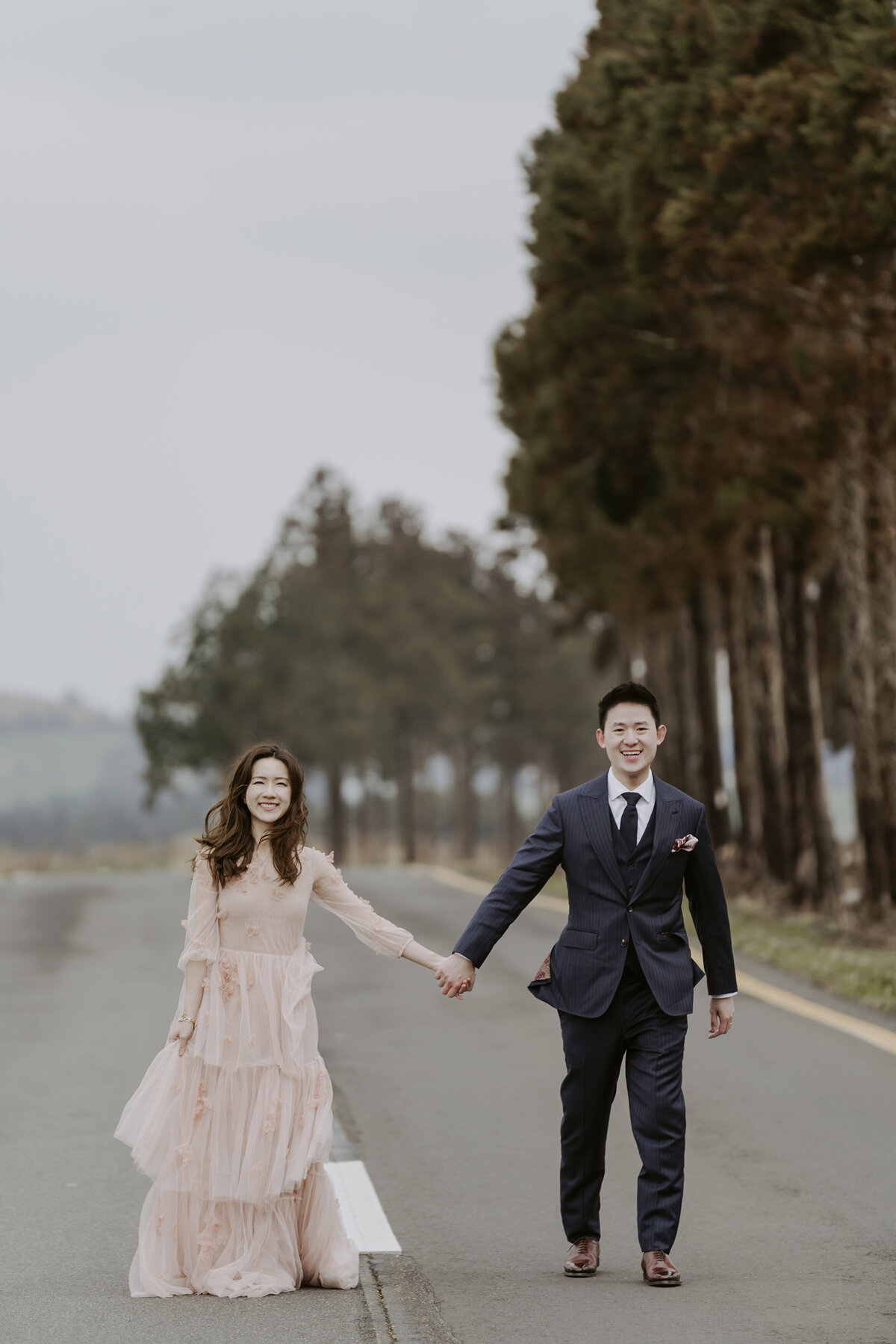 the bride wearing a raffled pink dress while the groom is wearing a black suit and tie while standing and holding hands in the road of jeju island