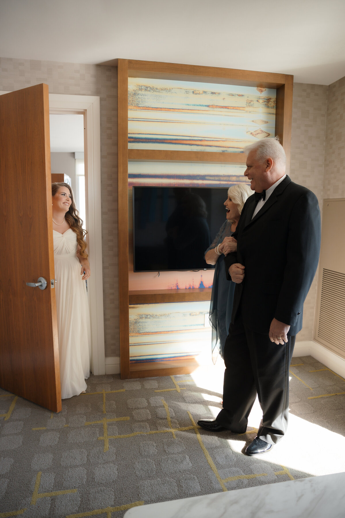 Bride sharing her first look with her family, who are smiling and cheering as they see her wedding gown for the first time in an elegant room.