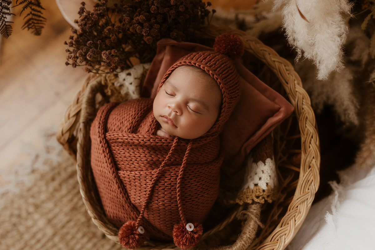 Newborn baby wrapped in a rust colored crochet blanket and matching hat sleeping in a basket.