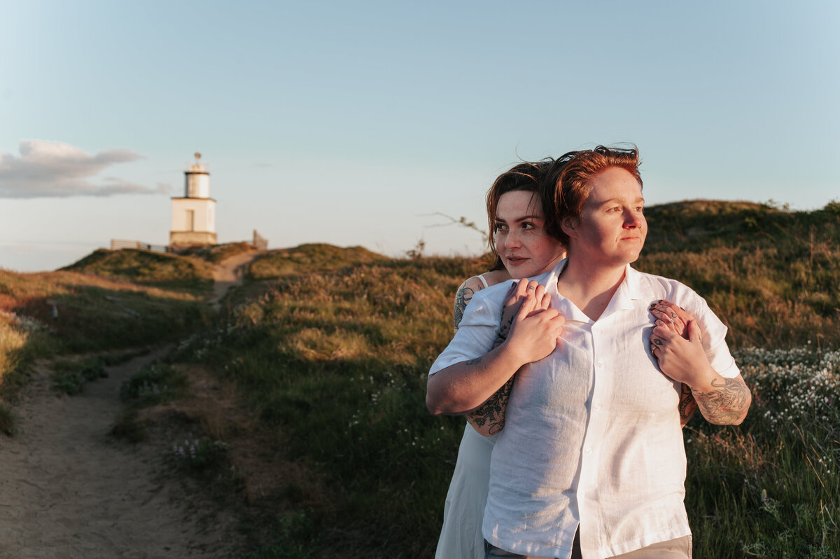 Romantic couple embracing on a coastal path with the iconic Cattle Point Lighthouse visible atop a hill in the San Juan Islands during golden hour, captured by a Pacific Northwest wedding photographer specializing in outdoor and destination elopements.