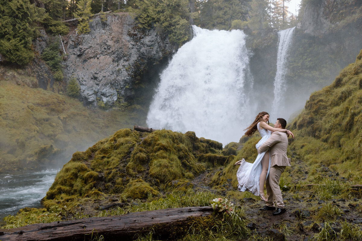 Groom holding up and swinging bride in front of a waterfall in Central Oregon.